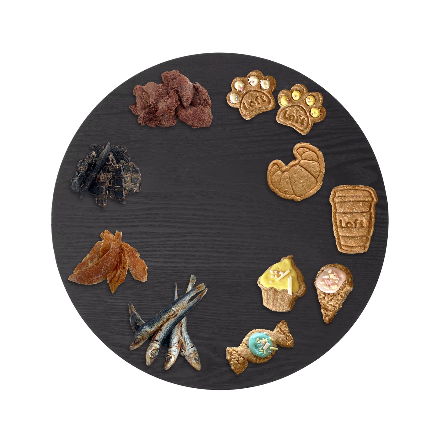 A round black serving platter displays an assortment of Laft Pet natural dog treats. On one half, gourmet natural treats: freeze-dried wild boar dehydrated lamb liver, chicken jerky, and pilchards are arranged. On the other half, high tea-inspired organic cookies: paw cookies, croissants, coffee cups, cupcakes, ice-cream cones, and lolly.
