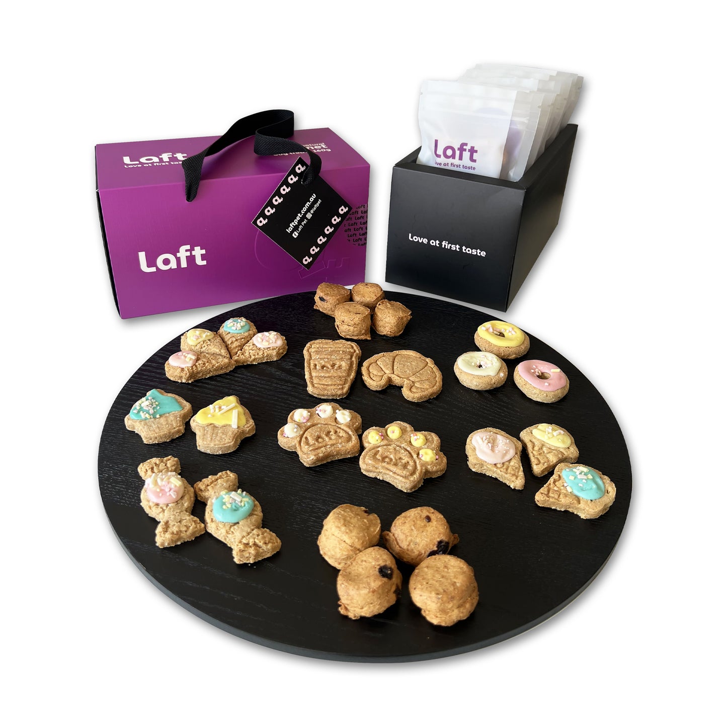 A presentation of the Laft Pet dog gift box. To the right, a sleek black container holds eight black-and-white pouches featuring the Laft logo and the tagline 'Love at first taste.' In the front, a round black serving platter displays various treats. The overall setup is clean, stylish, and visually appealing.