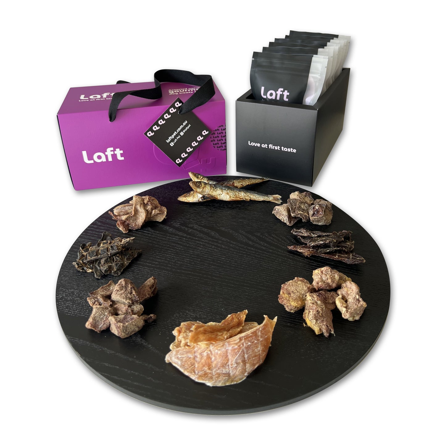 A presentation of the Laft Pet dog gift box. To the right, a sleek black container holds eight black-and-white pouches featuring the Laft logo and the tagline 'Love at first taste.' In the front, a round black serving platter displays various treats. The overall setup is clean, stylish, and visually appealing.