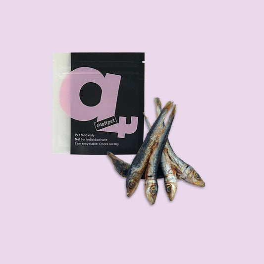 A black Laft Pet natural dog treat pouch placed next to a pile of dehydrated pilchards dog treats on a light purple background.