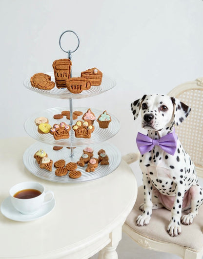 A Dalmatian wearing a purple bow tie sitting next to a three-tiered tray with Laft high tea dog biscuits and a cup of coffee on a white surface.