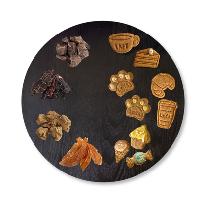 A round black serving platter displays an assortment of Laft Pet natural dog treats. On one half, gourmet natural treats: freeze-dried beef heart, kangaroo jerky, freeze-dried venison, and chicken jerky are arranged. On the other half, high tea-inspired organic cookies: cappuccino, cake slice, paw cookies, croissants, coffee cups, cupcakes, ice-cream cones, and lolly.