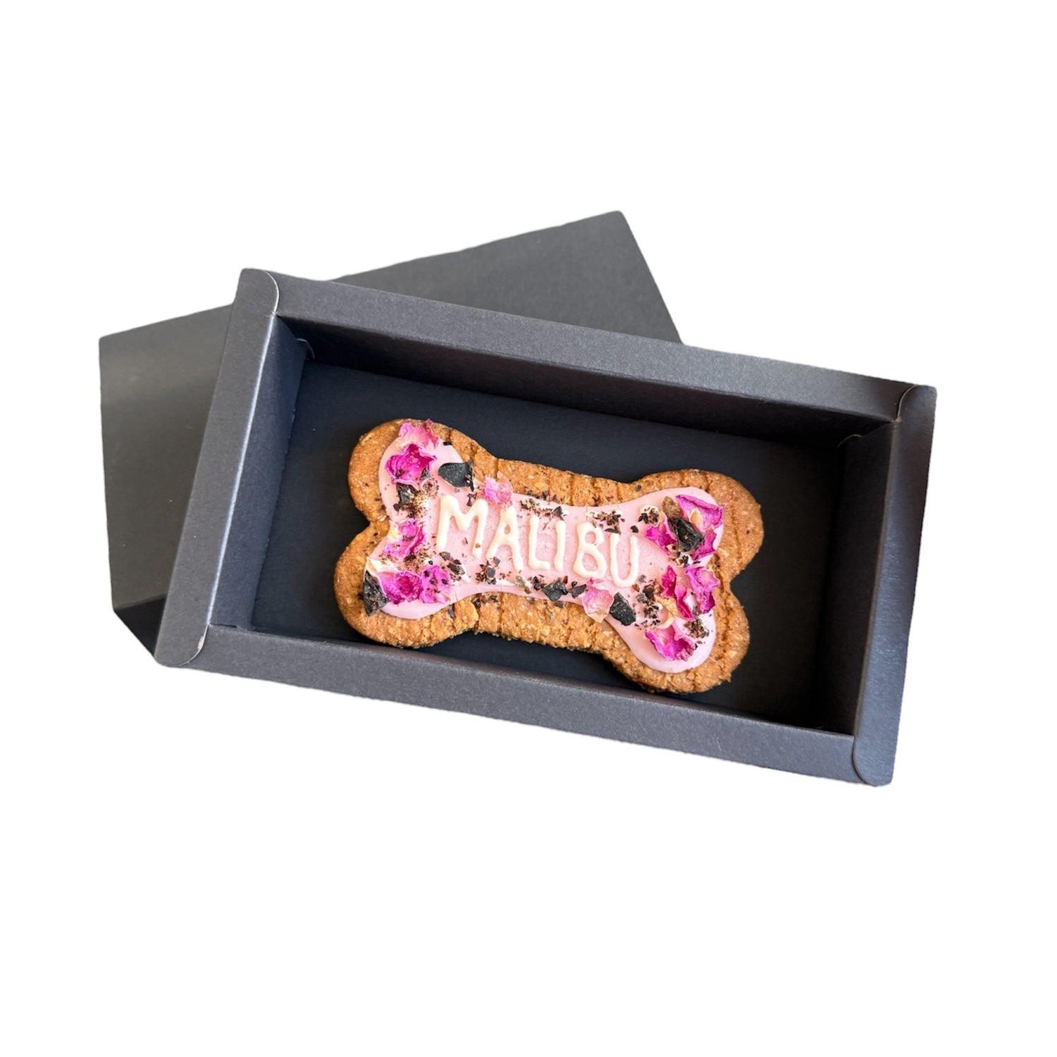 Personalised dog bone shaped biscuit, decorated with pink rose petals and pink icing with the name Malibu packed in a black gift box.