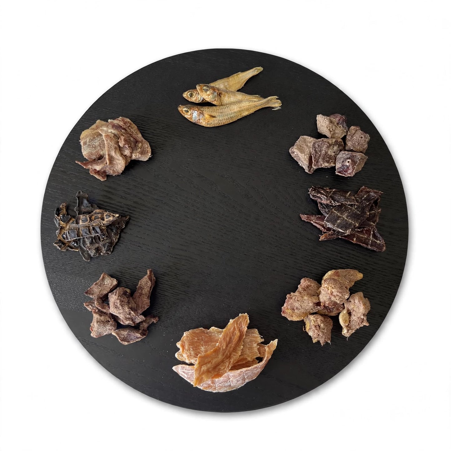 A round black serving platter displays various dehydrated and freeze-dried gourmet dog treats.
