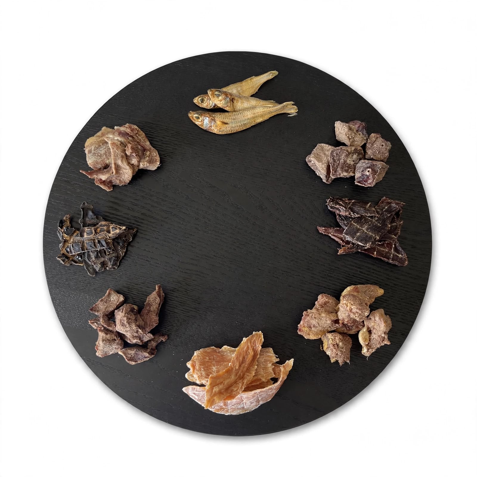 A round black serving platter displays various dehydrated and freeze-dried gourmet dog treats.

