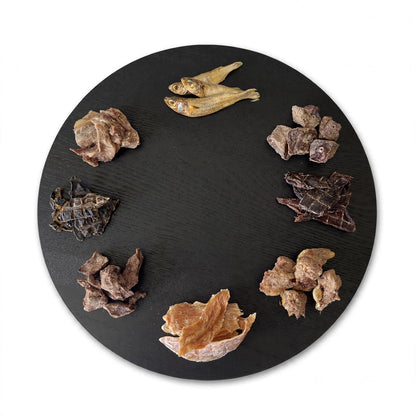 A round black serving platter displays various dehydrated and freeze-dried gourmet dog treats.
