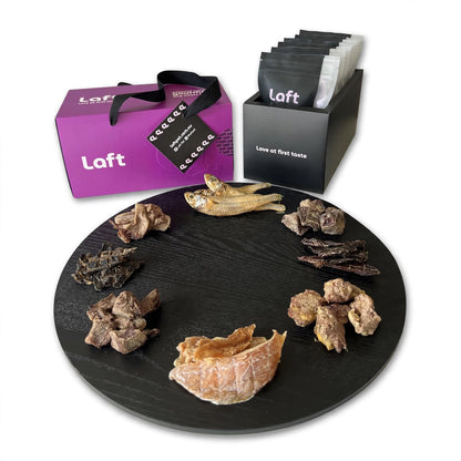 A presentation of the Laft Pet dog gift box. To the right, a sleek black container holds eight black-and-white pouches featuring the Laft logo and the tagline 'Love at first taste.' In the front, a round black serving platter displays various treats. The overall setup is clean, stylish, and visually appealing.