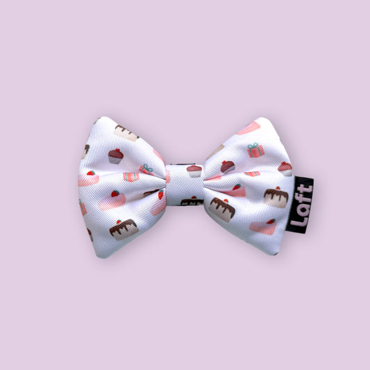 A white bow tie with dessert print and a Laft label on the right displayed on a light purple background.