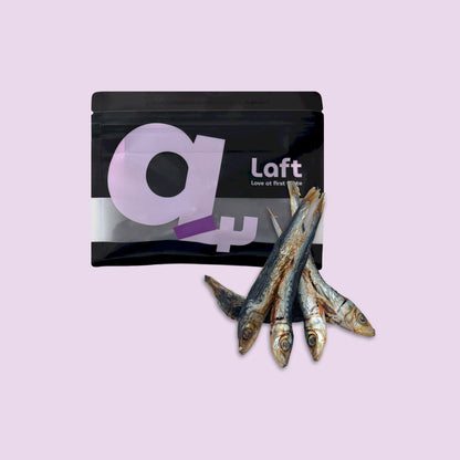 A black Laft Pet natural dog treat pouch placed next to a pile of dehydrated pilchards dog treats on a light purple background.